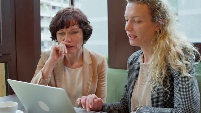 Two focused female colleagues having a business meeting in a cafe. The women are using a laptop, discussing a project, and mentoring
