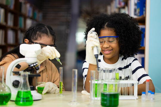 Group of diversity Young students explore science with curiosity and focus. Teacher guides them through microscope and green liquids. Books line shelves behind them, suggesting learning environment