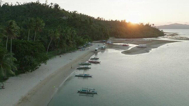 Aerial drone flight towards a golden sunset at Port Barton, Philippines. Traditional boats anchor in shallow water near a tranquil, palm-fringed beach. Perfect for peaceful travel promos.