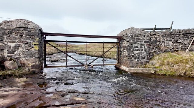 Rusty iron gate spanning a shallow stream at Burnhope Reservoir, framed by stone pillars and windswept moorland. slow motion