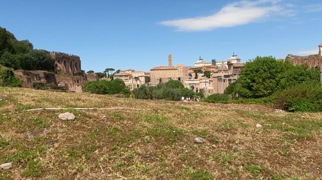 Sweeping ground-level view of the Roman Forum ruins in Rome, Italy. Ancient stone structures and historic buildings rise against a clear blue summer sky.