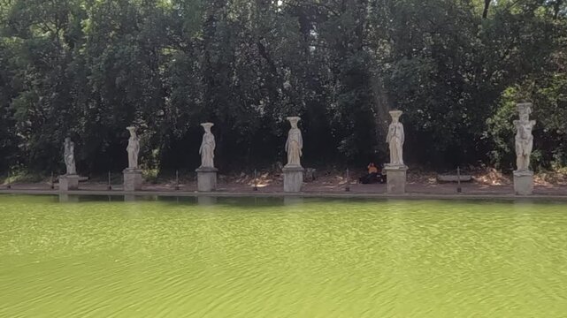 Row of classical caryatid statues lining the serene green water of Canopus pool at Villa Adriana, Tivoli near Rome. Ancient Roman heritage site surrounded by lush trees.