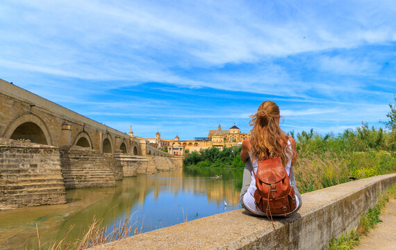 young female traveler sitting by the roman bridge in cordoba spain, solo traveler with leather backpack looking at the mosque-cathedral over guadalquivir river, authentic andalusian travel concept