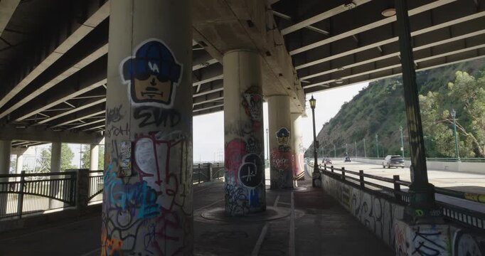 Cinematic daytime 4K 10-bit footage captured beneath the 5 and 110 freeway crossing in Los Angeles featuring large bridge support columns covered in distinct Los Angeles graffiti and street art.