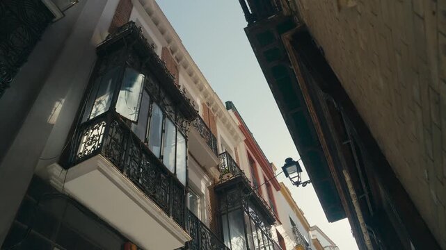 Low angle view of narrow street in Seville, Spain with historic facades, wrought iron balconies, and warm sunlight highlighting traditional architecture.