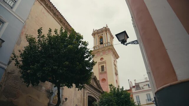Low angle view of a historic bell tower rising above narrow streets in Seville, Spain, framed by traditional buildings, street lamp, and greenery under soft daylight.