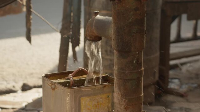 Hand pump filling metal container with flowing water on roadside, showing manual supply, daily use, public utility access, local street activity and basic needs