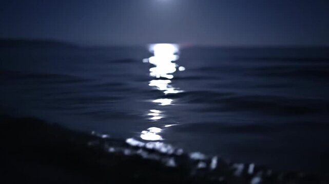 Bright moonlight creates a shimmering path on the dark, rippling surface of the ocean at night, offering a serene view.