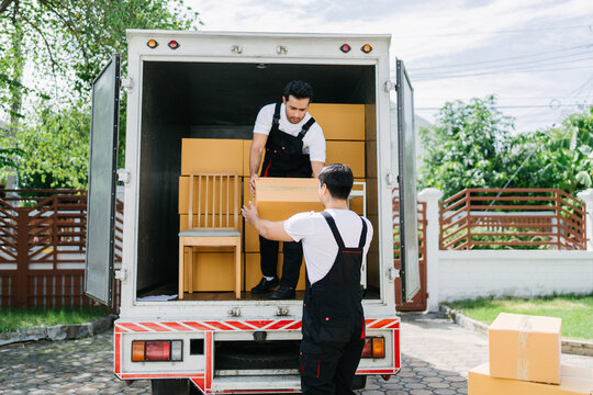Two male movers unloading cardboard boxes and furniture from a truck in a residential driveway. Professional moving service for home relocation and delivery. Efficient logistics for new home setup