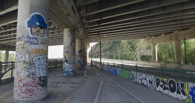 Cinematic daytime 4K 10-bit footage captured beneath the 5 and 110 freeway crossing in Los Angeles featuring large bridge support columns covered in distinct Los Angeles graffiti and street art.