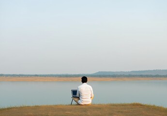 Fototapeta premium Man in casual clothing sitting on a grassy bank, using laptop with a serene lake and hills in the background