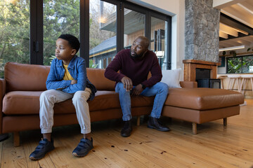 Naklejka premium African American father sitting with child on leather sofa at home, father leaning, child crossing