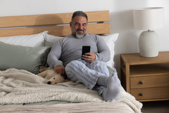 Mature man sitting on bed with headboard, holding smartphone and petting dog in striped pajamas