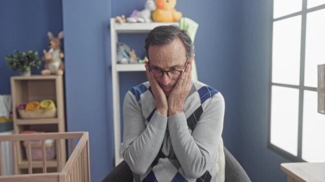 Man middle age hispanic clasping hands to mouth beside wooden crib and toy shelf in studio baby room with blue wall and window light; quiet concern.
