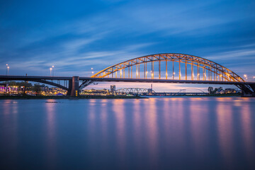 Fototapeta premium Nijmegen, a city in the netherlands on the waal river at night time during the blue hour with the lights on and reflections in the water. 