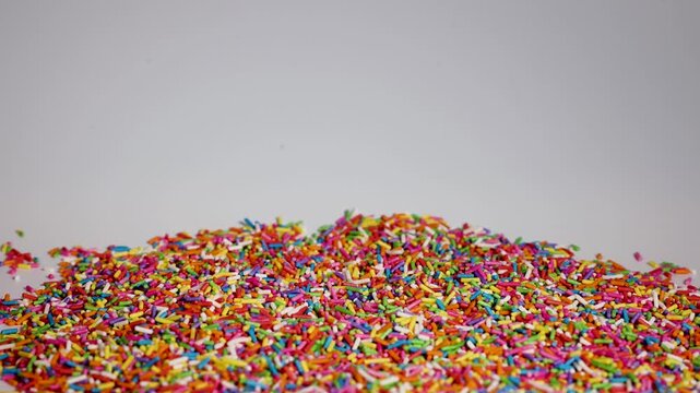 Colorful rainbow sprinkles falling on pile of sugar decorations in slow motion on white background.