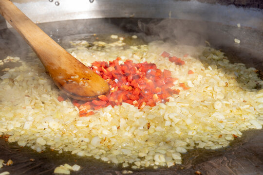 Detail of a sofrito of garlic and red pepper cooking in oil in a large paella pan. Close-up with a wooden spoon and steam, showing the traditional process of Mediterranean outdoor cooking.