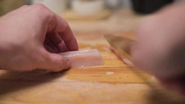 A person is cutting a chicken fillet on a wooden board in a kitchen. The individual appears to be focused and is using a knife to create even sections for cooking