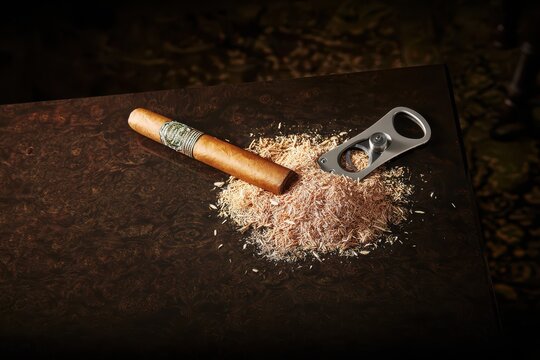 A premium cigar and cutter rest on a polished dark wood surface with tobacco shavings.