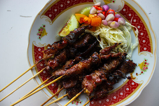 sate kambing or sate klatak or lamb satay or goat meat satay served with served with sliced ​​cabbage, ​​red onion, ​​chili, ​​tomatoes with soy sauce on plate. isolated white background -
