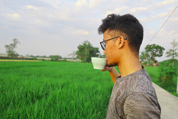 Fototapeta premium A man is drinking coffee in a field. The field is green and the sky is blue. The man is wearing a gray shirt and glasses
