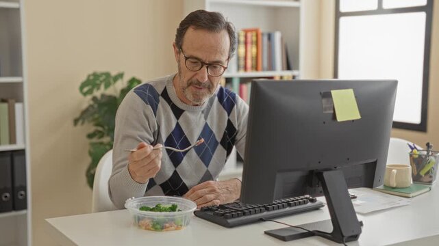 Middle aged man eating salad with fork at desk in building, wearing glasses and argyle sweater while using computer; concentration healthy lunch.