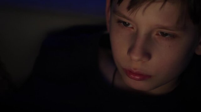 Close-up of a boy's face illuminated by the flickering light of a monitor in a dark room. A teenager sits in front of a computer screen at night