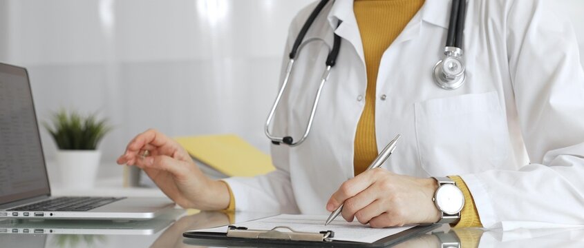 Doctor wearing stethoscope and yellow sweater under white lab coat, making notes on a clipboard in a bright medical office. Medicine, healthcare and science concept