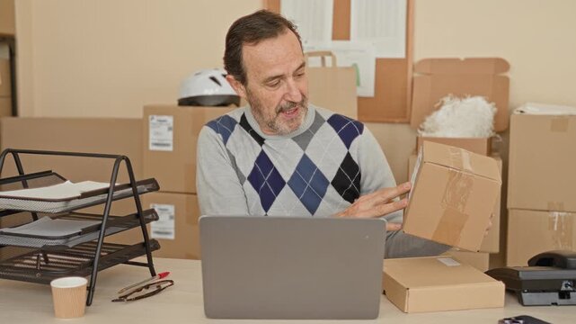 Man holding a cardboard box while tapping a laptop keyboard in a building office parcel workspace; small business focus.