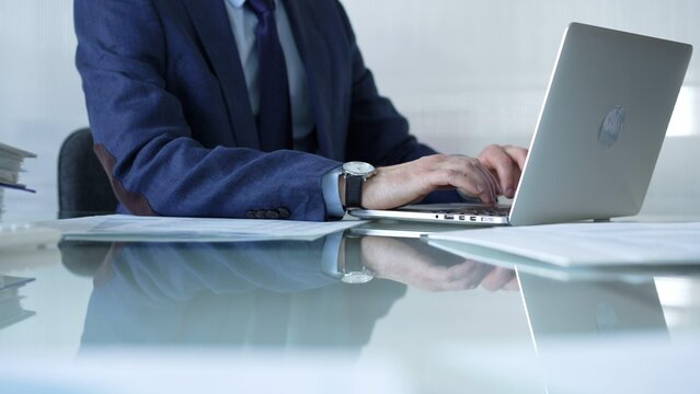 Businessman in blue suit is typing on laptop at glass desk while sitting in bright office, showcasing productivity and modern work environment. Businesspeople concept