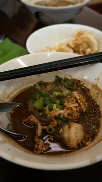 Traditional Thai boat noodles with thick spicy pork broth, meatballs, and liver in ceramic bowl with chopsticks