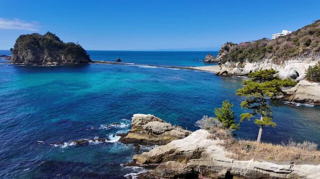 A sweeping high-angle pan across the crystal-clear turquoise waters of Dogashima, Japan. The shot captures the rugged volcanic cliffs, a lone pine tree, and the iconic Sanshiro Islands