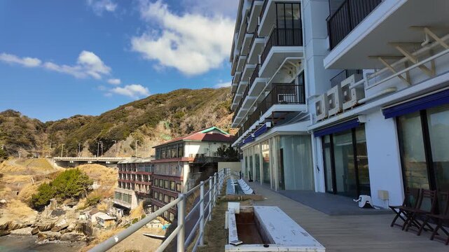The camera pans down from a modern white seaside hotel and across a sunlit terrace to reveal a stunning panoramic view of Dogashima's turquoise waters