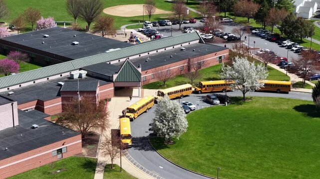 Top-down aerial view of U.S. elementary school during spring with yellow school buses lined up for student pickup, green lawns and sunny midday light in typical American suburban setting. Approach.