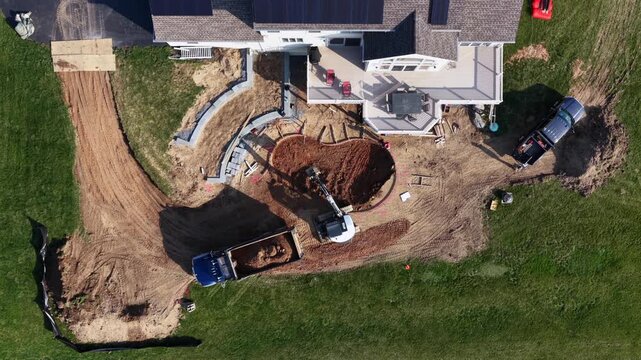 Top-down aerial view of suburban American home with workers excavating backyard swimming pool, excavator and dump truck moving soil near raised deck and landscaped yard in sunny daylight.