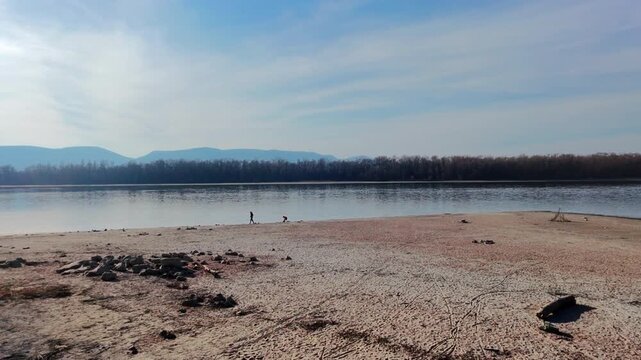 A professional dolly-in drone shot glides toward two small silhouettes walking on the vast, sandy shore of the Danube. The scene features the calm river and hazy blue mountains of Sződliget under a cl