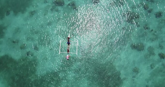 Aerial top view drone shot descending over a traditional Filipino outrigger boat with children rowing in clear turquoise water in Carabao Island near Boracay Philippines