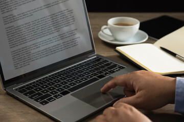 Fototapeta premium Hand using laptop trackpad with text document beside notebook, tablet, and coffee cup on desk, representing content writing, copy editing, digital publishing, article review, and remote workflow produ