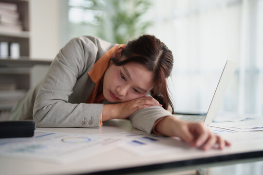 Stressed asian female office worker experiencing burnout, feeling tired and sad, sleeping on desk from overwork and exhaustion during long hours in corporate environment