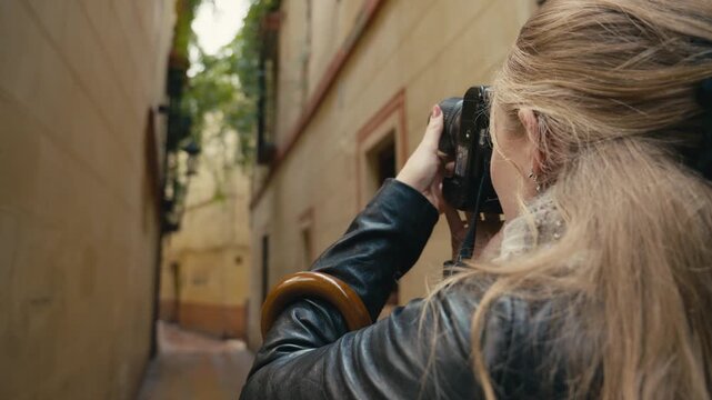 Rear view of a female tourist taking photos with a camera in a narrow street in Seville, Spain, surrounded by historic buildings and soft natural light, capturing travel and exploration.
