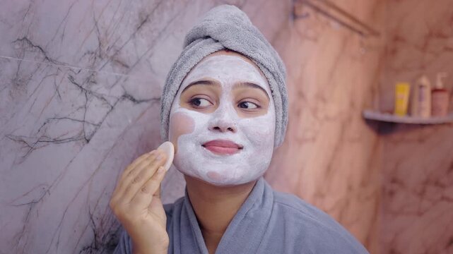 Female model carefully cleansing facial mask residue using a cotton pad during her skincare routine indoor.