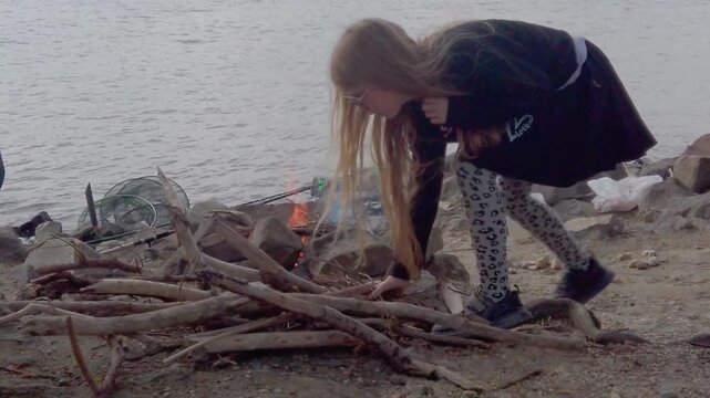 A young girl in patterned leggings carefully adds hay or dry wood to a small, rocky campfire on the Danube riverbank. Fishing rods and a net are visible in the background against the gentle movement o