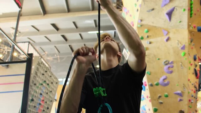 Male athlete carefully pulls safety rope for insurance. Trainer works in gym lifting client up to climbing wall slow motion