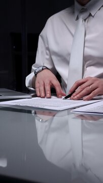 Man in white shirt reviewing legal documents, checking details of a contract with a magnifying glass on a dimly lit desk. Audit, business and overworking concepts
