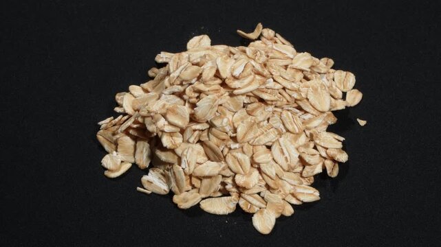 Macro of organic rolled oats pile rotating on black background, food texture.