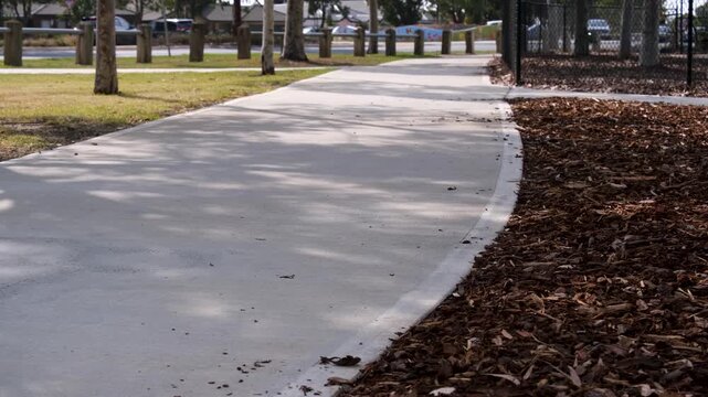A concrete pedestrian footpath under dappled tree shade runs alongside a busy suburban road with passing traffic, flanked by a woodchip bark mulch garden bed. Active travel infrastructure in Australia