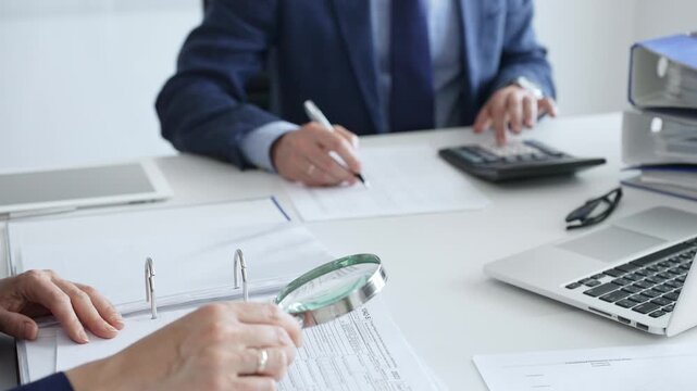 Business colleagues are working together, analyzing financial documents with a magnifying glass, calculating and taking notes in the office, focus is on the woman hands. Audit and taxes in business