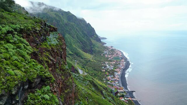 A breathtaking aerial view of Paul do Mar, Madeira, where verdant cliffs meet the Atlantic, showcasing a charming village nestled along the dark pebble beach under a cloudy sky.