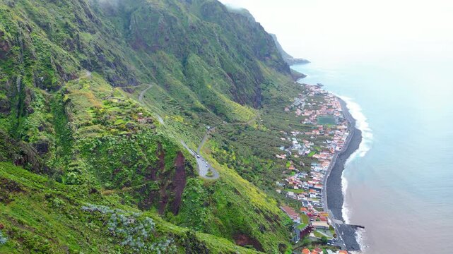 A breathtaking aerial view of Paul do Mar, Madeira, where verdant cliffs meet the Atlantic, showcasing a charming village nestled along the dark pebble beach under a cloudy sky.