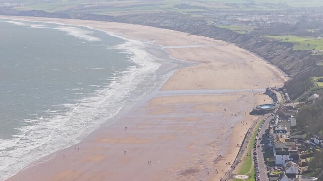 Filey Bay in Filey North Yorkshire England with Ravine Road seafront, wide intertidal sandy beach, promenade and tidal pool along North Sea coastline beneath eroding cliffs, drone slow pan left shot